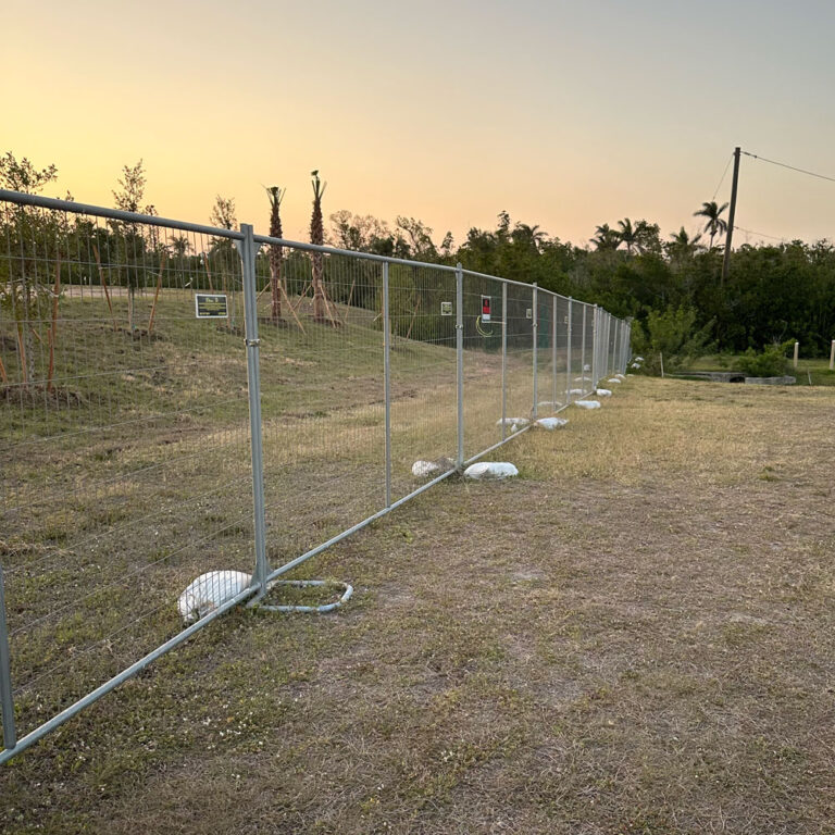 Freestanding temporary fence panels used for event or property security at sunset, supported by sandbags on a grassy lot.